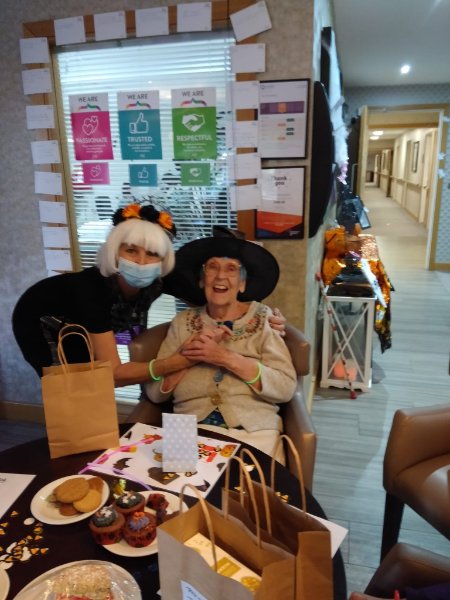 Two women at a Halloween party: one woman with a white wig, and an elderly woman sitting happily on a chair wearing a witch hat.