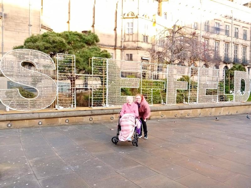 resident and carer in front of a big Sheffield sign in sheffield town centre smiling
